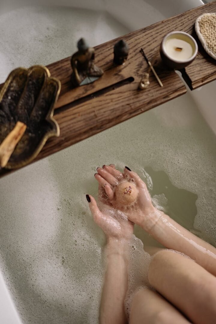 5 ideas para convertir tu rutina de baño en un spa en casa 3 Serene bath setting featuring hands holding a bath bomb, surrounded by candles and a wooden tray.