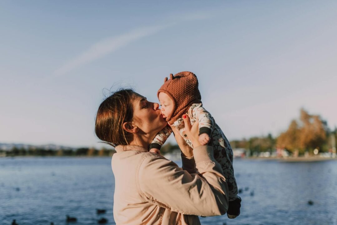 A woman lovingly kisses a baby by the lake during a sunny autumn day.
